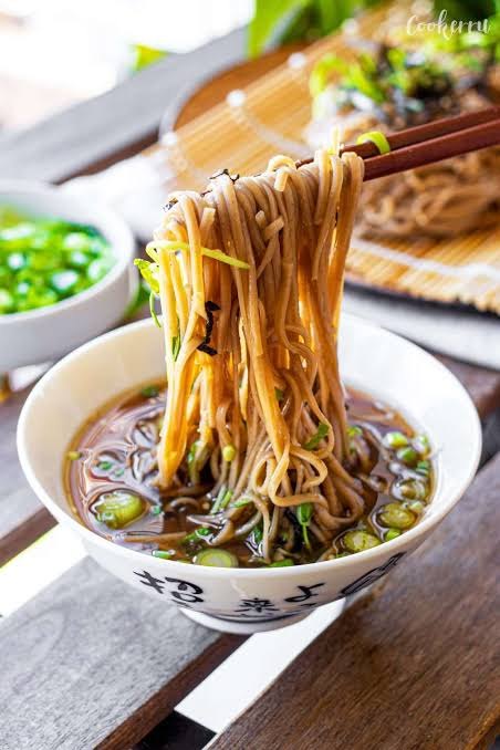 Boekweitnoedels met stokjes opgetild uit een kom dipsaus, met zaru soba op de achtergrond
