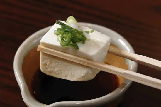 Een blokje zachte tofu tussen stokjes, boven een schaaltje saus met lente-ui
